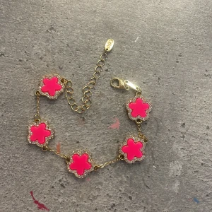Guldarmband med rosa blommor - Armband i guldton med fem blommor i starkt rosa, omgivna av små glittrande stenar. Blommorna har en tydlig form och armbandet har justerbar kedja med karbinlås. Perfekt för dig som vill sticka ut med färg och bling.