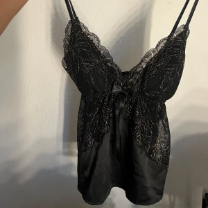 Svart spetslinnetopp med rosett - Supersöt svart linnetopp med tunna axelband, spetsdetaljer och broderade blommor i guld. Toppen har en liten rosett framtill och är gjord i glansigt satinliknande material. Perfekt för dig som gillar en feminin och elegant stil.