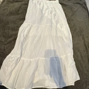 Vit långkjol med volanger från Lindex - Säljer en vit långkjol från Lindex med volangdetaljer och resår i midjan. Kjolen är tillverkad i mjuk bomull och har en luftig, flowy känsla som passar perfekt för varma dagar. Enkel och stilren design som är lätt att matcha med olika toppar.