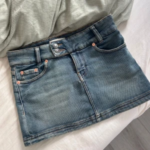 Blå jeanskjol - Blå lågmidjad minikjol i klassisk modell med fem fickor och dubbla knappar framtill. Kjolen är kort och har en tight passform. Har aldrig använt.