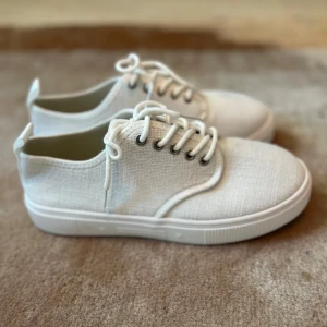 Vita sneakers i textil, storlek 39 - Snygga vita sneakers i textil med klassisk design och rund tå. Skorna har vita snören, silverfärgade öljetter och en platt sula i gummi. Perfekta för dig som gillar en clean och stilren look till vardags. Lätta och bekväma att bära.