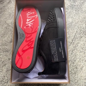 Christian Louboutin svarta sneakers med nitar - Säljer ett par svarta sneakers från Christian Louboutin med ikonisk röd sula och coola svarta nitar på tån. Skorna är i mocka och skinn, har snörning och en platt sula. Perfekta för dig som vill sticka ut med lyxig streetstyle.