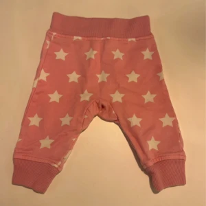 Rosa babybyxor med stjärnor, storlek 56 - Supersöta rosa babybyxor från Kappahl i storlek 56. Byxorna har vita stjärnor över hela tyget och breda ribbade muddar i midja och benslut. Mjukt bomullsmaterial som är skönt mot huden. Perfekta för små barn som vill vara både bekväma och stiliga.