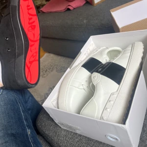 Valentino Garavani Open sneakers vit/svart - Säljer ett par snygga Valentino Garavani Open sneakers i vitt skinn med bred svart läderdetalj över vristen. Skorna har klassisk snörning, rund tå och ikoniska nitar längs sulan. Perfekta för dig som vill ha en clean men lyxig look. Låda och allting följer med