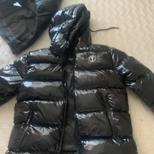 Svart glansig pufferjacka Moose Knuckles - Säljer en svart glansig pufferjacka från Moose Knuckles med huva och dragkedja. Jackan har två fickor med dragkedja och tydlig logga på bröstet. Perfekt för kalla dagar när du vill hålla stilen.