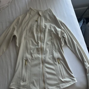 Vit träningsjacka - Säljer en beige träningsjacka med snygga gulddetaljer på dragkedjor och logga. Jackan har figurnära passform, hel dragkedja framtill och två fickor med dragkedja.