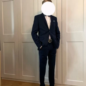 Mörkblå kostym med kavaj och byxor - Säljer en stilren mörkblå kostym med matchande kavaj och byxor och skjortan. Kavajen har klassisk passform, enkla knappar och fickor framtill. Byxorna är raka och har bälte med metallspänne. Perfekt för dig som vill vara extra snygg på speciella tillfällen. Endast använd enstaka gånger!