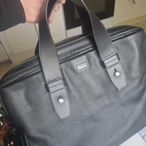 Svart portfölj i skinn från BOSS - Snygg svart portfölj från BOSS i äkta skinn med stilrena handtag och silverfärgade detaljer. Väskan har en klassisk, rektangulär form och dragkedja upptill. Perfekt för laptop och viktiga papper, med diskret BOSS-logga framtill och matchande bag tag.