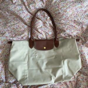 Beige Longchamp axelväska med skinn - Snygg beige axelväska från Longchamp med bruna detaljer i skinn och guldknapp framtill. Väskan har klassisk form, långa handtag och är tillverkad i slitstarkt syntetmaterial med skinnöverdrag på handtag och lock.