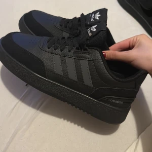 Svarta Adidas sneakers i skinn - Snygga svarta Adidas sneakers med klassiska tre ränder på sidan och Adidas-logga på plösen. Skorna är tillverkade i skinn med detaljer i syntet och har snörning samt rund tå. Perfekta för dig som gillar en clean och stilren look.
