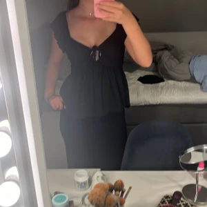 Svart blus med volang och knyt - Säljer en svart blus med djup v-ringning, volangdetaljer vid ärmarna och knyt framtill. Toppen har en lös passform nedtill och är ärmlös med breda axelband. Perfekt för dig som gillar en feminin och trendig stil.