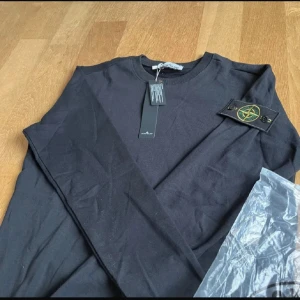 Svart långärmad tröja Stone Island - Svart långärmad tröja från Stone Island med klassisk patch på ärmen. Tröjan har rund halsringning och ribbade muddar vid ärmslut och nederkant. Tillverkad i mjuk bomull, perfekt för en clean och stilren look.