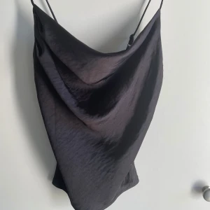 Svart linnetopp - Säljer en svart linnetopp med tunna justerbara axelband och snygg draperad front. Toppen har en slät, glansig yta som ger en lyxig känsla och faller snyggt över kroppen. Perfekt för dig som gillar minimalistisk och stilren design.