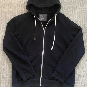 Svart hoodie med dragkedja från H&M - Svart hoodie från H&M med vit dragsko och hel dragkedja framtill. Klassisk modell med huva, kängurufickor och ribbade muddar. Bra skick, storlek M passar TTS, vid fler frågor eller funderingar tveka inte på att skriva!