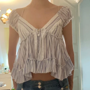 Randig blus från Kimchi Blue S - Supersöt blus från Kimchi Blue i vitt och ljuslila med tunna ränder. Modellen är croppad med volanger, spetsdetaljer och knappar framtill. Blusen har korta ärmar, v-ringning och öppen rygg med knytband. 