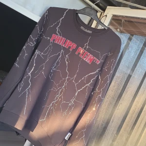 Svart Philipp Plein tröja med blixtmönster - Svart långärmad tröja från Philipp Plein med glittrande blixtmönster i silver och röd logga framtill i stenar. Tröjan har rund halsringning och en snygg detalj på ryggen. Perfekt för dig som vill sticka ut med en cool och exklusiv look. Storleken tar vi i privat.