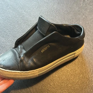 Svarta sneakers från Axel Arigato - Svarta slip-on sneakers från Axel Arigato i skinn med vit sula och diskret guldtext på sidan. Klassisk rund tå och platt sula, stilren design utan snörning. Perfekta för dig som gillar minimalistisk och clean look.
