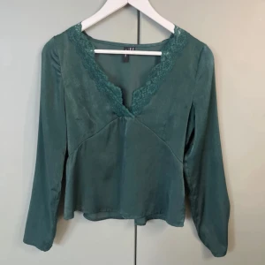 Grön blus med spets från Vero Moda - Supersnygg grön blus från Vero Moda med v-ringning och spetsdetaljer längs halsen. Blusen har långa ärmar och en lätt glansig yta som ger en lyxig känsla. Resår i midjan baktill för en smickrande passform. 