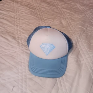 Blå och vit truckerkeps med diamant - Snygg truckerkeps i ljusblått och vitt med ett snyggt diamanttryck. Kepsen har mesh-bakstycke för extra ventilation och justerbar snapback för perfekt passform. Perfekt för sommaren. Använd fåtal gånger