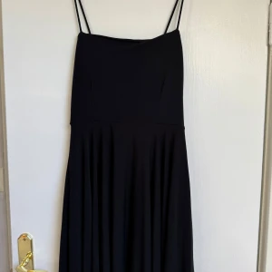 Svart långklänning med smala axelband - Svart långklänning med tunna axelband och stilren siluett. Klänningen har en mjukt fallande kjol och korsade band i ryggen som ger en snygg detalj. Perfekt för dig som gillar minimalistisk och elegant stil. Materialet känns lätt och följsamt mot kroppen.