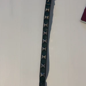 Smalt grönt  nitararmband i skinn valentino - Snyggt smalt grönt armband i skinn med silvriga fyrkantiga nitar längs hela bandet. Perfekt accessoar för att ge din outfit en edgy vibe. Klassisk metallspänne och justerbara hål för bästa passform.