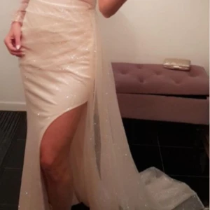 Glittrig champagnefärgad långklänning - Supersnygg långklänning i champagnefärg med glittrigt tyg och hög slits. Klänningen har en elegant och figurnära passform, perfekt för att sticka ut. Tunn, svepande material som ger en lyxig känsla.