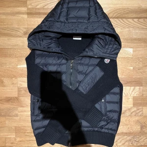 Moncler Cardigan - Säljer en snygg svart cardigan från Moncler. Den har en quiltad design med dragkedja och långa ärmar i ribbat material. Perfekt för höst och vinter, håller dig varm och stilren. Jackan har en huva och är i bra skick. Passar både till vardags och lite finare tillfällen.