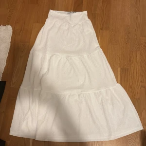 Vit långkjol med volanger - Säljer en vit långkjol med flera volanglager. Kjolen har en luftig och flowy känsla, perfekt för varmare dagar. Den är enkel och stilren, vilket gör den lätt att matcha med olika toppar.