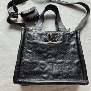 Svart axelväska Juicy Couture - Cool svart axelväska från Juicy Couture i syntetmaterial med präglat logomönster och hjärtan. Väskan har bred axelrem med logga, JC-metallogo framtill och en liten avtagbar ficka. Invändigt foder med tryckt logga och dragkedja.