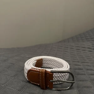 Vitt flätat bälte med bruna skinn-detaljer - Snyggt vitt flätat bälte med bruna skinn-detaljer och silvrigt spänne. Bältet har en stilren look med tydliga sömmar på skinnet och passar perfekt till jeans eller chinos. Flätad design gör det enkelt att justera längden efter behov.