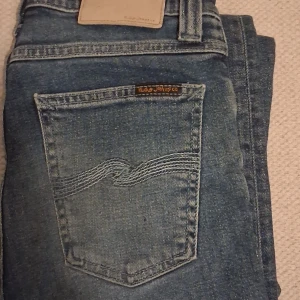 Nudie Jeans Co - Säljer ett par klassiska blå jeans från Nudie Jeans Co med snygga detaljer på bakfickan och logga ovanför. Jeansen har normal passform, fem fickor och är tillverkade i slitstarkt denim. Perfekta för dig som gillar stilrena och tidlösa jeans. Storlek w28.