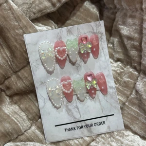 Rosa och vita press-on naglar med pärlor - Supergulliga press-on naglar i rosa och vitt med 3D-dekorationer som pärlor, hjärtan och stora glittriga rosetter. Naglarna har en mix av glansig finish och transparenta detaljer, perfekta för en söt och unik look.