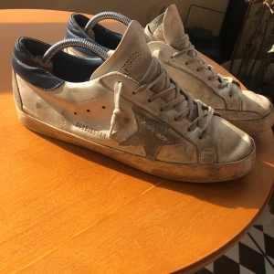 Golden Goose Superstar sneakers vit/blå - Golden Goose Superstar sneakers i vitt skinn med grå mockadetaljer och den ikoniska stjärnan på sidan. Mörkblå hälkappa i skinn, snörning framtill och platt sula. Snyggt slitna för en cool, avslappnad look. Perfekta för dig som gillar streetstyle.