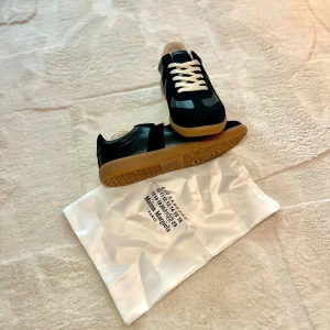 Maison Margiela svarta sneakers - Svarta sneakers från Maison Margiela med beige sula och vita snören. Skorna har en klassisk, stilren design med inslag av mocka och skinn. Perfekta för dig som gillar minimalistisk och trendig stil.