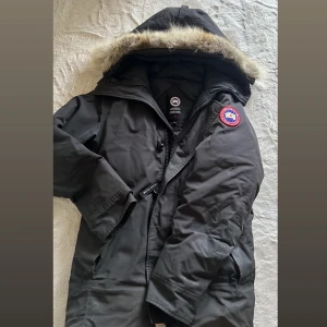 Canada Goose - Säljer en svart parkas från Canada Goose med ikonisk patch på ärmen och äkta päls runt huvan. Jackan har dragkedja, knappar och flera fickor. Perfekt för kalla vinterdagar och har en klassisk, stilren look. Pris går att diskutera 