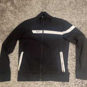 Svart ziptröja från BOSS med vita detaljer - Säljer en svart långärmad ziptröja från BOSS med vita detaljer på bröstet och ena ärmen. Tröjan har hel dragkedja framtill och två fickor med vita dragkedjor. Perfekt för en clean och sportig look.