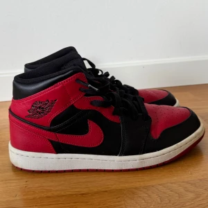 Nike Air Jordan 1 Mid Svart/Röd - Säljer ett par Nike Air Jordan 1 Mid i klassisk svart och röd färg. Skorna har en snygg kontrast mellan svart och rött skinn, med svart snörning och vit sula. Ikoniska Jordan-loggan på sidan och tungan. Perfekta för dig som gillar streetwear och sneakers med attityd.