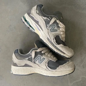 New Balance 2002R Protection Pack grå - Säljer ett par New Balance 2002R Protection Pack i grå och beige. Skorna har en chunky sula, slitna mockadetaljer och meshpaneler för en rå och trendig look. Ikoniskt N på sidan och breda snören. Perfekta för dig som gillar streetwear och snygga sneakers.