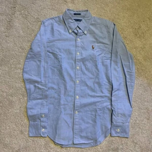 Blå skjorta från Ralph Lauren - Klassisk ljusblå skjorta från Ralph Lauren med button-down krage och broderad logga på bröstet. Skjortan har lång ärm, vita knappar och är tillverkad i bomull. Perfekt för dig som gillar stilren och tidlös design.