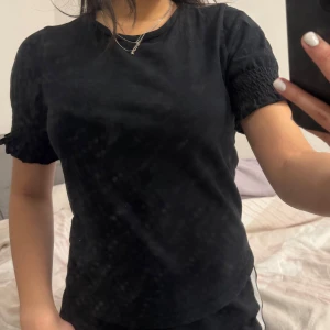 Svart t-shirt med puffärm och smock - Svart t-shirt med korta puffärmar och smockdetalj vid ärmslutet. T-shirten har rund halsringning och är gjord i mjuk bomull. Enkel men ändå trendig med volangkant på ärmarna för en extra touch.