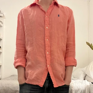 Rosa linneskjorta Polo Ralph Lauren - 174 på bilden och väger 56kg. Snygg rosa skjorta från Polo Ralph Lauren i relaxed fit. Skjortan är tillverkad i linne, har långa ärmar, klassisk krage och knäppning framtill. Broderad blå logga på bröstet ger en clean och stilren look.
