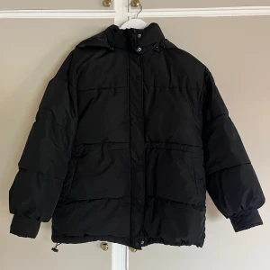 Svart pufferjacka med huva - Svart pufferjacka med huva och stora fickor. Jackan har dragkedja och knappar framtill, samt elastiska muddar vid ärmsluten. Perfekt för kalla dagar med sin oversized passform och quiltade paneler. Snygg och enkel att matcha till allt.