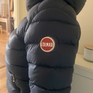 Svart Colmar pufferjacka med huva - Säljer en svart pufferjacka från Colmar med stor huva och tydlig logga på ärmen. Jackan har dragkedja och är perfekt för kalla dagar. Den är vadderad och har en modern, sportig look som passar dig som vill hålla stilen även när det är kallt.