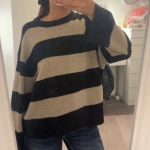 Randig oversized stickad tröja - Säljer en oversized stickad tröja med breda svarta och beige ränder. Tröjan har lång ärm och en avslappnad passform, perfekt för dig som gillar en chill och trendig stil. Supermjuk och skön att bära till jeans eller leggings.