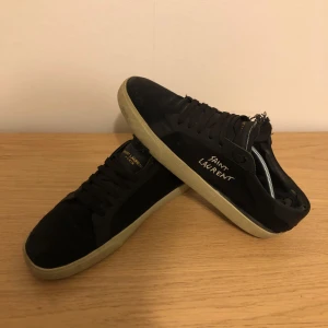 Saint Laurent Court Classic - Svarta sneakers från Saint Laurent med broderad logga på sidan och gulddetaljer på plösen. Skorna har en klassisk låg profil, snörning och beige sula. Materialet är mocka och skinn, vilket ger en lyxig känsla och snygg kontrast.