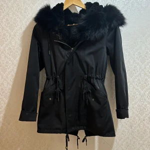 vinterjacka - Svart parkajacka med stor huva och bred fuskpäls runt kragen och insidan. Jackan har dragsko i midjan, stora fickor med knappar och dragkedja framtill. Perfekt för kalla dagar och ger en riktigt snygg siluett.