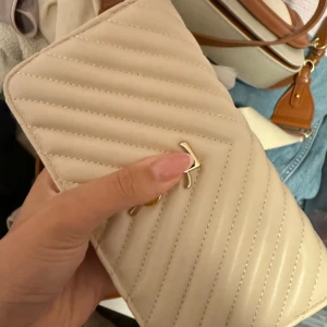 Beige quiltad plånbok med dragkedja - Snygg beige plånbok med quiltad design och guldfärgad dragkedja. Plånboken har flera fack för kort och mynt, samt ett stilrent metallspänne på framsidan. Perfekt storlek för att ha med sig överallt och passar till de flesta stilar.YZL märke 