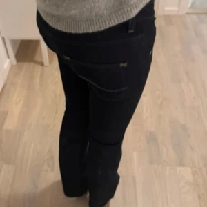 Mörkblå bootcut jeans från Lee - Säljer ett par mörkblå jeans från Lee med bootcut passform och låg midja. Jeansen har dubbla knappar framtill, klassiska fickor och snygga detaljer med kontrastsömmar. Materialet är jeans och de har en dragkedja. Perfekta för dig som gillar en tidlös look.