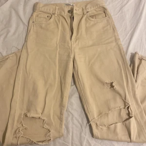 Beige ripped jeans med raka ben - Beiga jeans från Pull&Bear med raka ben och flera slitningar på både fram- och baksida. Byxorna har klassisk femficksdesign, råa kanter vid benslut och är tillverkade i bomull. Perfekt för dig som gillar en avslappnad och trendig look.