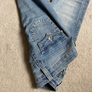 True Religion Joey Low Rise Flare Jeans - Snygga ljusblå jeans från True Religion, modell Joey Low Rise Flare. Bilderna är tagna innan de tvättas så de kommer inte vara smutsiga vid foten. Storlek: 24 och de har inte så mycket tecken på användning bortom benen. Hör av er vid frågor och funderingar!🥰
